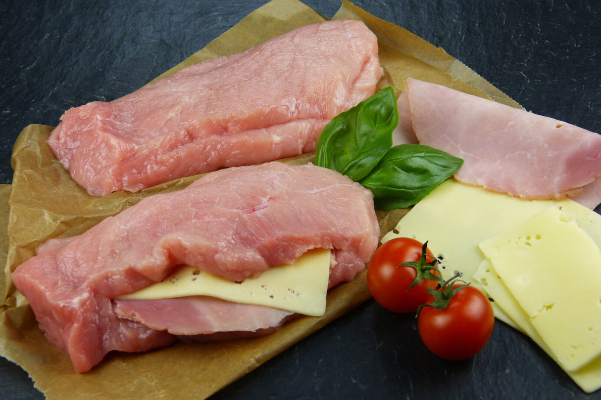 Eine leere Schweine Tasche für Cordon Bleu und eine mit Käse und Kochschinken gefüllt angerichtet auf Backpapier mit Tomaten Käse Basilikum und Kochschinken auf Schieferhintergrund