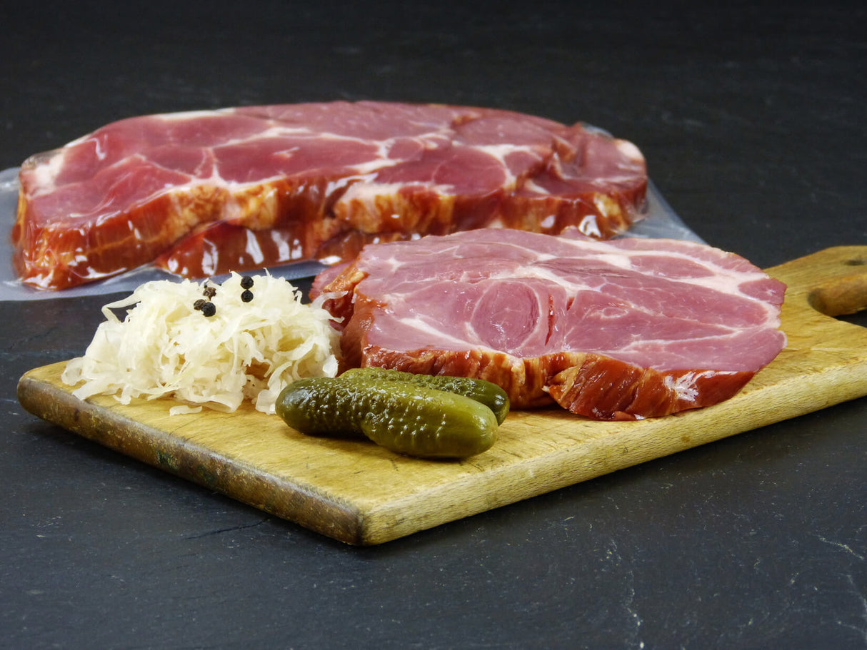 Kasseler Nackenbraten verpackt und eine Scheibe Kasseler Nackenbraten angerichtet mit Sauerkraut Pfefferkörnern und zwei Gewürzgurken auf einem Holzschneidebrett mit Schieferhintergrund