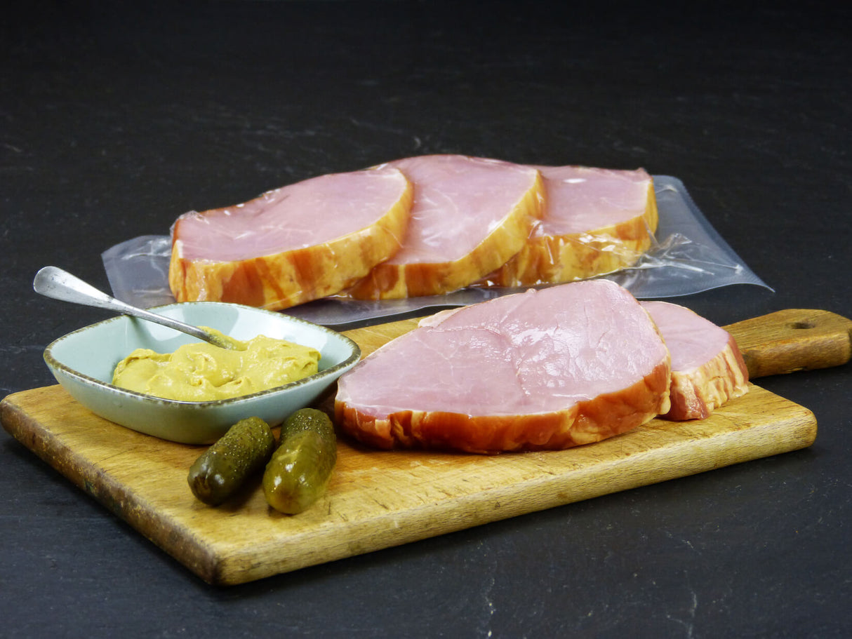 Kasseler Lachsbraten verpackt und angerichtet auf einem Holzschneidebrett mit zwei Gewürzgurken Senf in einer Schüssel und einem Löffel drinne auf einem Holzschneidebrett mit Schieferhintergrund