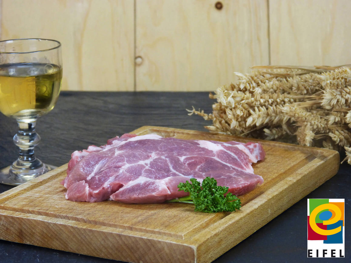 Kammsteak vom EIFEL Schwein angerichtet mit Petersilie Weizen und einem Weinglas auf einem Holzschneidebrett mit einem Schieferholzhintergrund im Vordergrund rechts das Regionalmarke EIFEL Logo