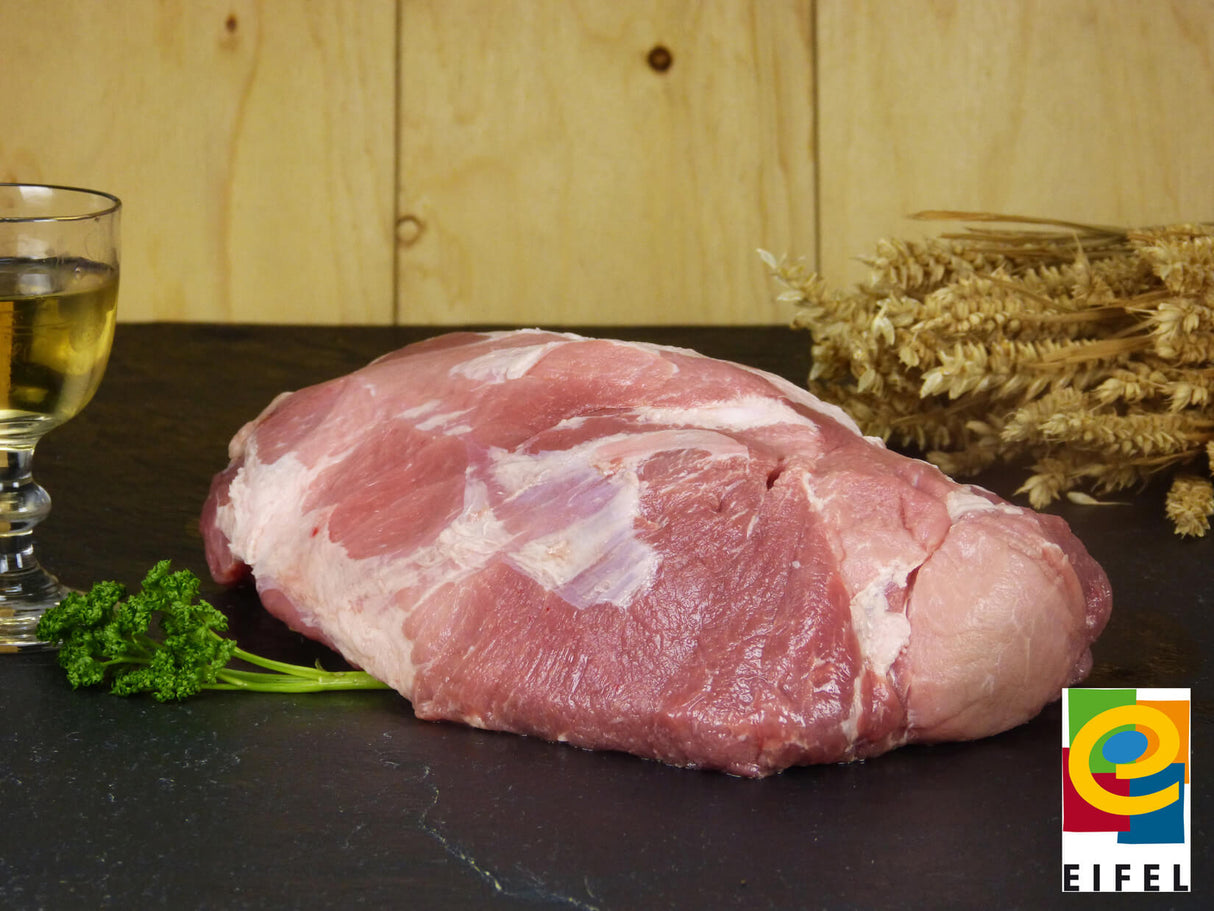 Kamm ohne Knochen vom EIFEL Schwein angerichtet mit Petersilie Weizen und einem Weinglas auf einem Schieferholzhintergund im Vordergrund rechts das Regionalmarke EIFEL Logo