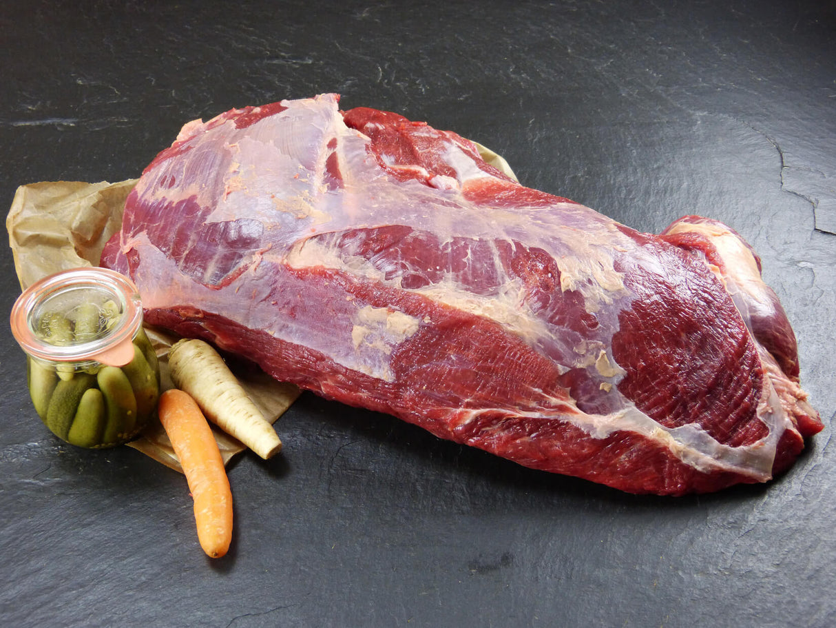 Dickes Bugstück vom EIFEL Rind angerichtet mit Sellerie einer Karotte und einem Gewürzgurkenglas auf Backpapier mit Schieferhintergrund