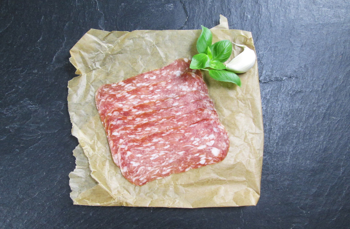 Salami Spianata Romana in scheiben geschnitten angerichtet mit Basilikum und einer Knoblauchzähe auf Backpapier mit Schieferhintergrund