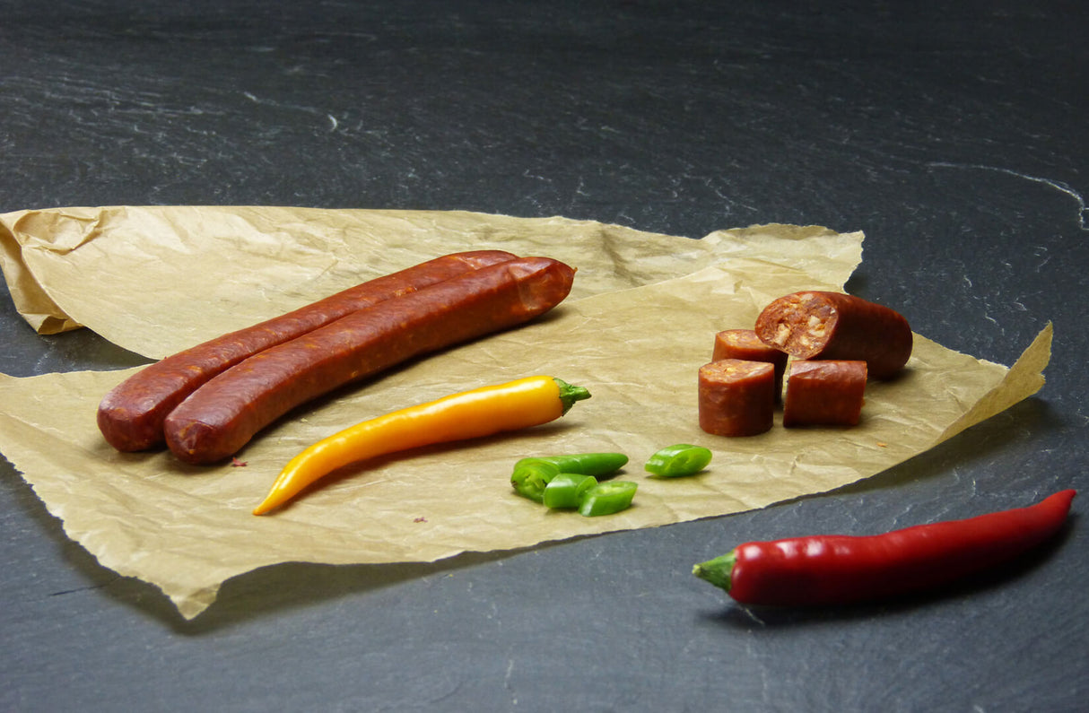Zwei Chili Schinkenmettwürstchen vom EIFEL Schwein angerichtet mit einer gelben Chilischote und einer roten dazu kommen grüne stücke von einer Chilischote und vier stücke einer Chili Schinkenmettwurst auf einem Backpapier mit Schieferhintergrund