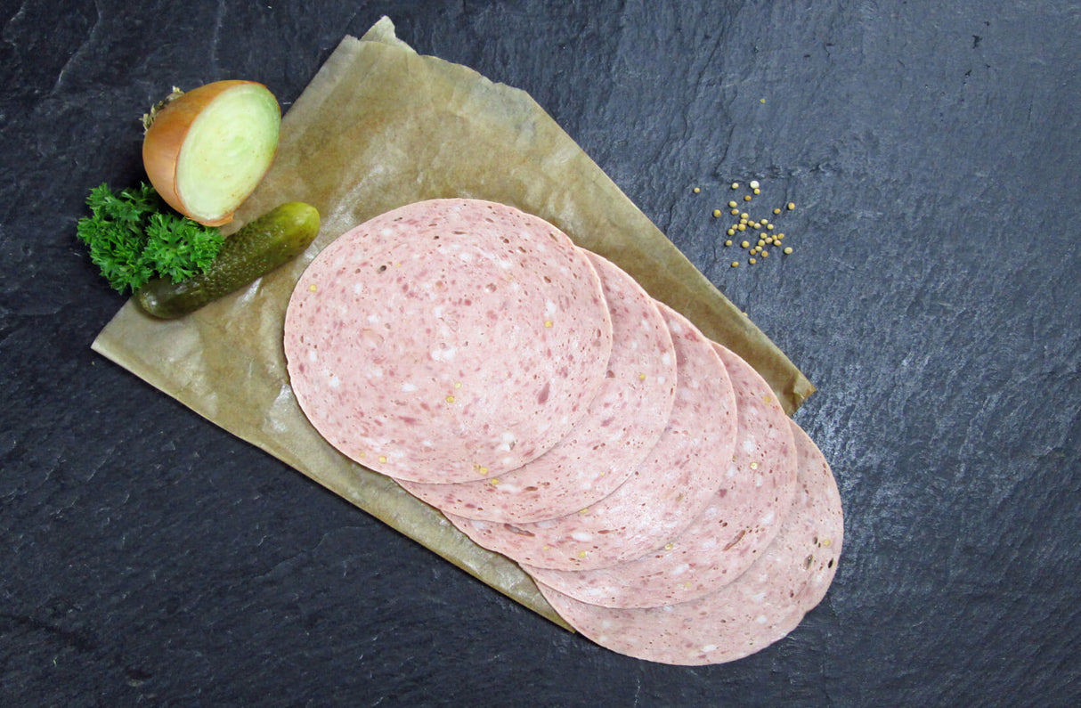 Fünf scheiben Bierwurst vom EIFEL Schwein angerichtet mit einer halben Zwiebel Petersilie Senfkörnern und einer Gewürzgurke auf Backpapier mit Schieferhintergrund