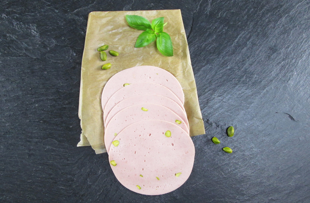 Fünf scheiben Mortadella vom EIFEL Schwein angerichtet mit Pistazien und Basilikum auf backpapier mit Schieferhintergrund