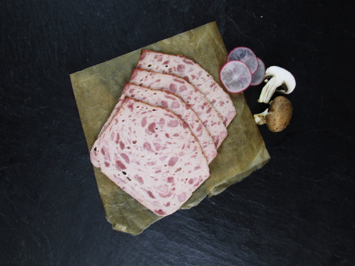 Vier scheiben Fleischkäse grob gebacken vom EIFEL Schwein angerichtet mit 3 halben Radieschen und einem Halbierten Chmpignon Pilz auf Backpapier mit Schieferhintergrund