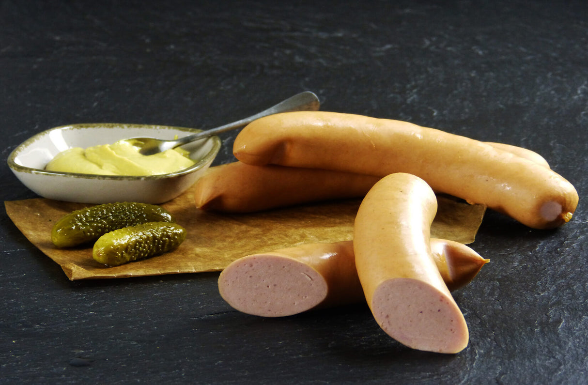 Zwei Fleischwürstchen vom EIFEL Schwein mit einem halbierten Fleischwürstchen angerichtet mit zwei Gewürzgurken und Senf in einer kleinen Schüssel mit einem Löffel drinne auf Backpapier mit Schieferhintergrund