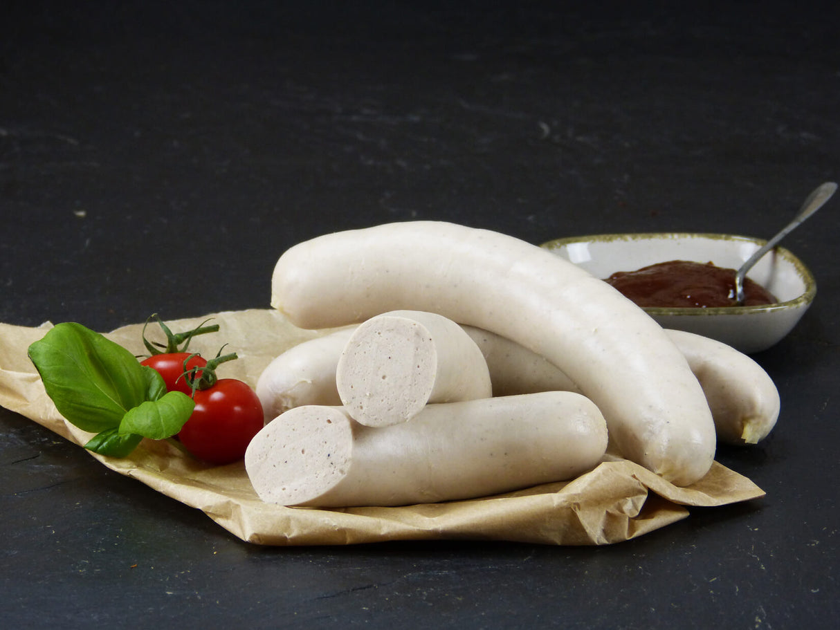 Zwei Bratwürste vom EIFEL Schwein und eine halbierte Bratwurst angerichtet mit Ketchup in einer Schüssel und einem Löffel drinne zudem zwei Tomatetn und Basilikum auf Backpapier mit Schieferhintergrund