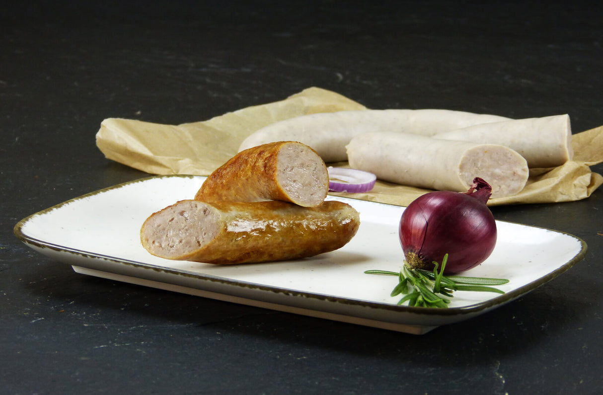 Eine halbe grobe gegrillte Bratwurst angerichtet auf einem Teller mit einer roten Zwiebel Thymian und mit einer gebrühten Bratwurst und eine halbierte gebrühte Bratwurst angerichtet mit zwei roten Zwiebelringen auf Backpapier mit Schieferhintergrund