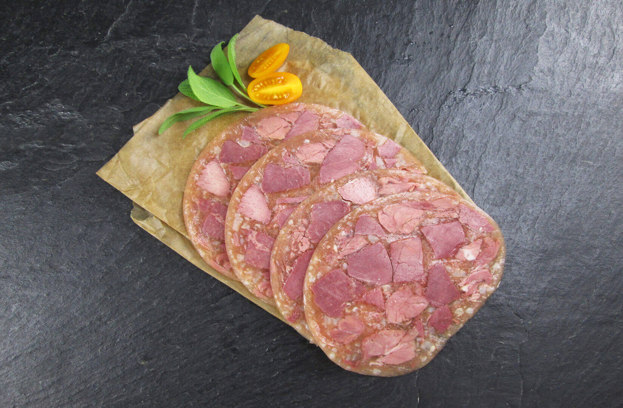 Vier scheiben Schwartenmagen mit Rindfleisch angerichtet mit einer halbierten gelben kleinen Tomate und Minze auf Backpapier mit Schieferhintergrund