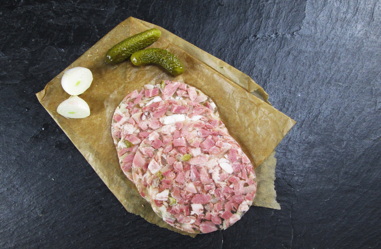 Vier scheiben Schweinskopfsülze mit Gurken angerichtet mit einer halbierten Zwiebel und zwei Gewürzgurken auf Backpapier mit Schieferhintergrund