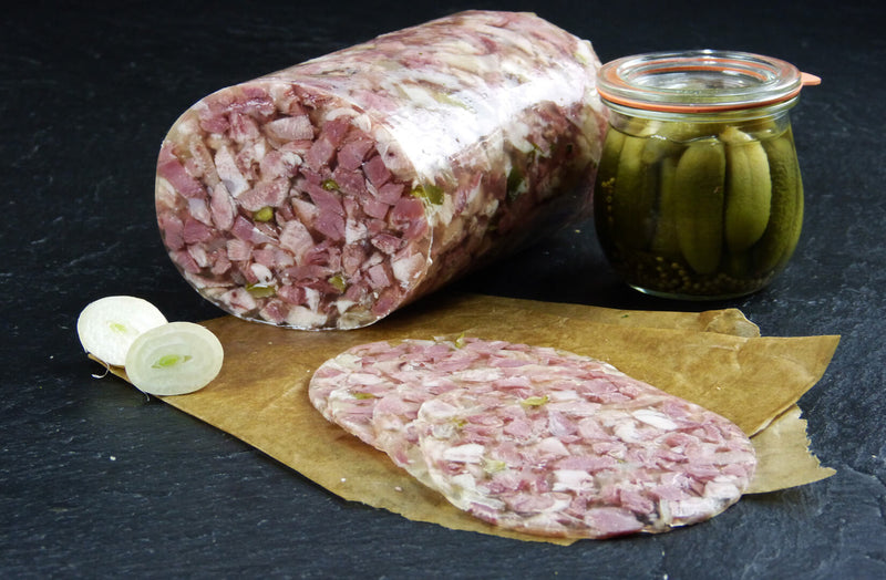 Ein Anschnitt Schweinskopfsülze mit Gurken und vier scheiben Schweinskopfsülze angerichtet mit einer halbierten Zwiebel und eine Gewürzgurkenglas auf Backpapier mit Schieferhintergrund