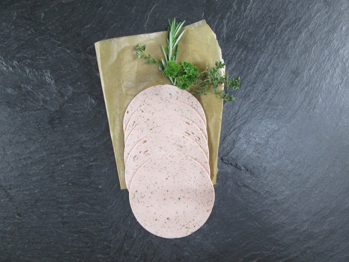 Fünf scheiben Kräuterlyoner vom EIFEL Schwein angerichtet mit Thymian Rosmarin und Petersilie auf Backpapier mit Schieferhintergrund