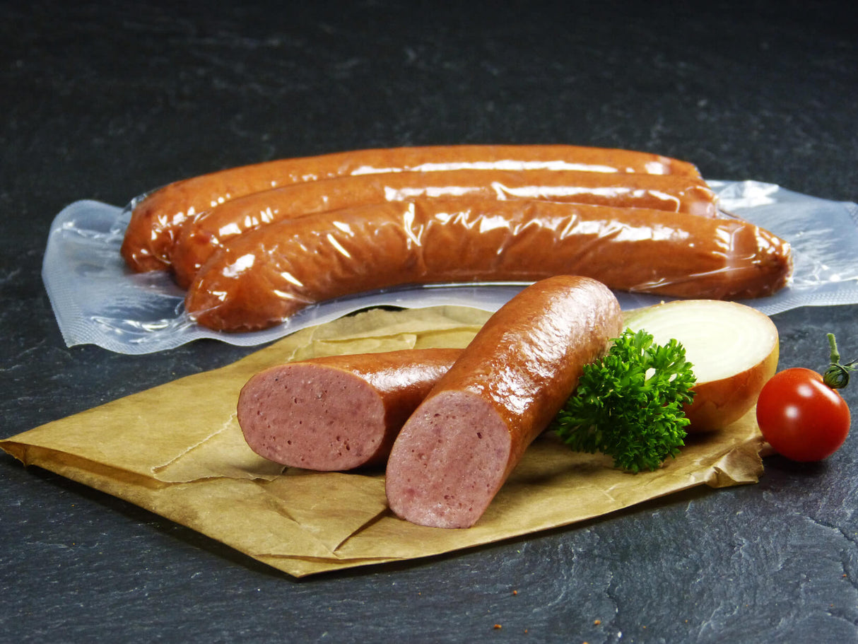 Drei Rindswürstchen vom EIFEL Rind verpackt und ein halbiertes Rindswürstchen angerichtet mit Petersilie einer halben Zwiebel und einer Cherrytomate auf Backpapier mit Schieferhintergrund