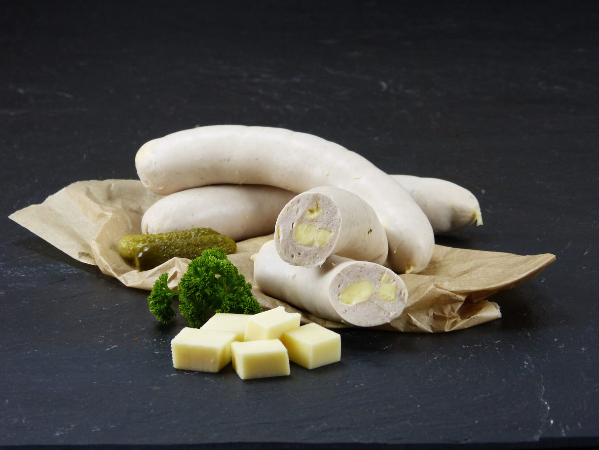 Zwei Käsebratwürste vom EIFEL Schwein und eine halbierte Käsebratwurst angerichtet mit Petersilie Gewürzgurken und ein paar Käsewürfeln auf Backpapier mit Schieferhintergrund