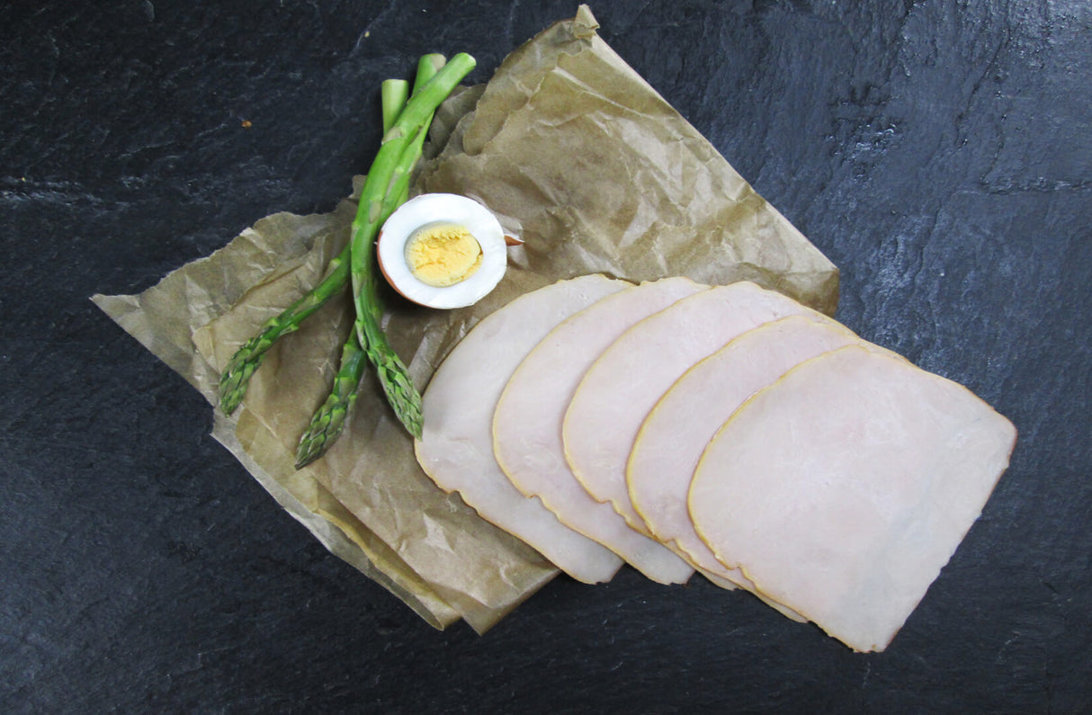 Fünf scheiben Putenbrust geräuchert angerichtet mit grünem Spargel und einem halben Ei auf Backpapier mit Schieferhintergrund