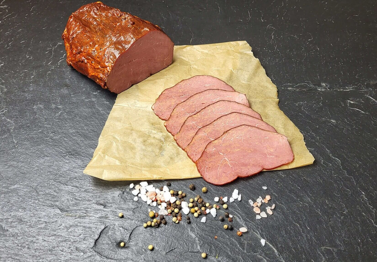 Pastrami am Stück und in Scheiben geschnitten dekoriert mit Pfefferkörner und Salz auf Schieferhintergrund