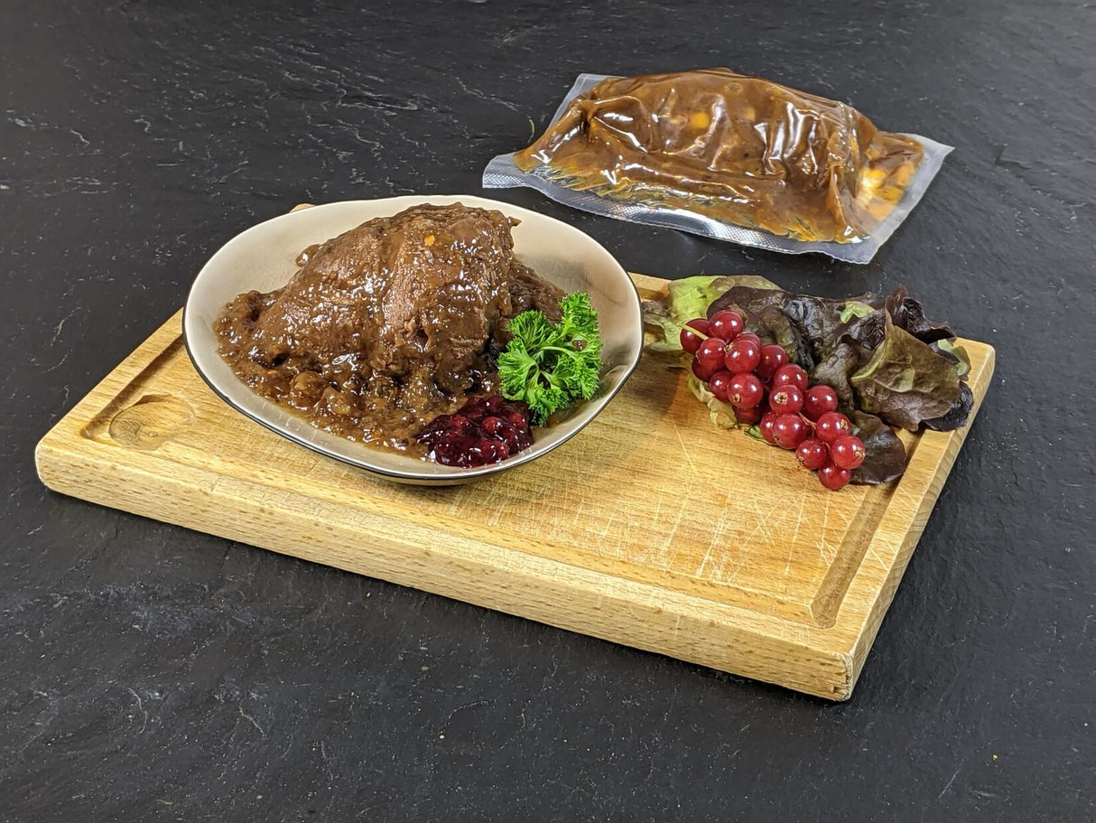 Rinderbäckchen gegart in Sauce im Kochbeutel und gegart auf Teller auf Holzbrett mit Johanniesbeeren und Salatdeko auf Schieferhintergrund