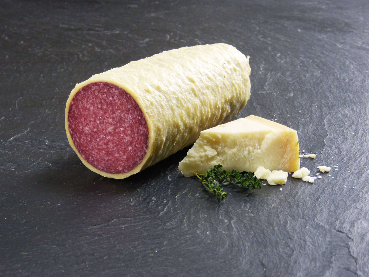 Salami im Käsemantel am Stück auf Backpapier mit Parmesankäse und Kräutern auf Schieferhintergrund