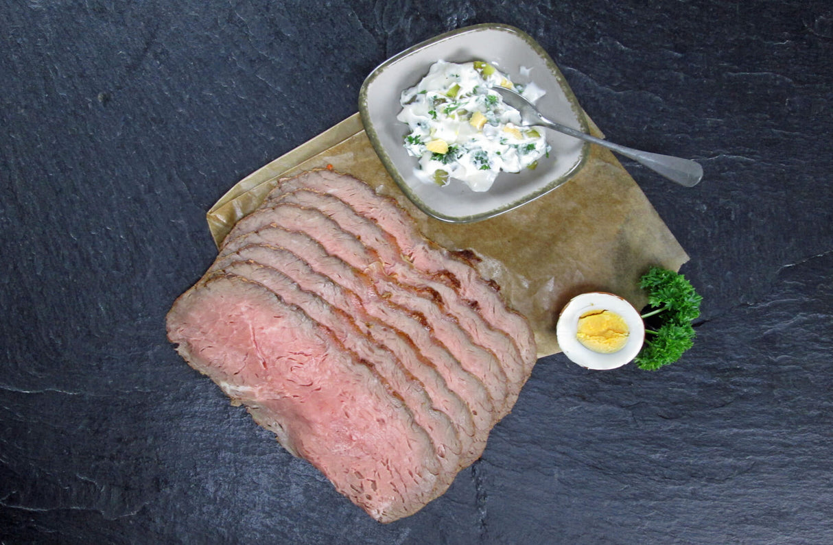 Roastbeefbraten geschnittenin Scheiben auf Backpapier mit Ei und Petersilie und einer Schale Sauce auf Schieferhintergrund