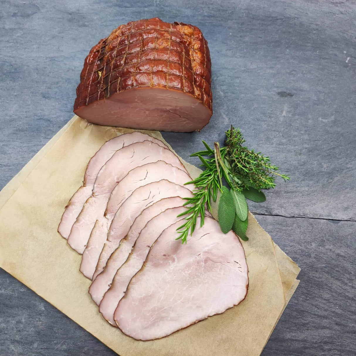 Kesselschinken vom EIFEL Schwein am Stück und 7 Scheiben aufgeschnitten mit verschiedenen Kräutern auf Holzpapier und Schieferhintergrund