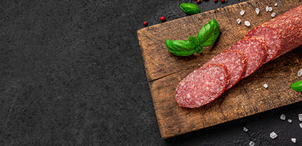 Salami in Scheiben geschnitten mit Basilikum und groben Pfefferkörnern auf Holzbrett mit Schieferhintergrund