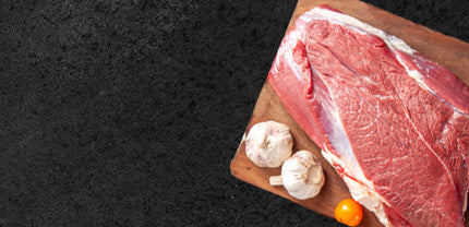 Rohes Fleischstück vom Eifelrind auf Holzbrett dekoriert mit Knoblauch und einer kleinen gelben Tomate auf schwarzem Hintergrund