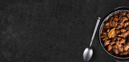 Rinder Gulasch in Gußpfanne gegart mit Löffel auf Schieferhintergrund