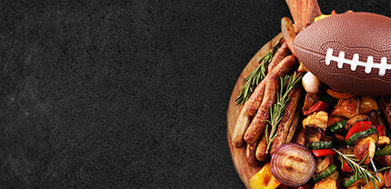 Holzbrett mit einem Football Rosmarien Bratwürstchen Zwiebeln und Paprika auf Schieferhintergrund