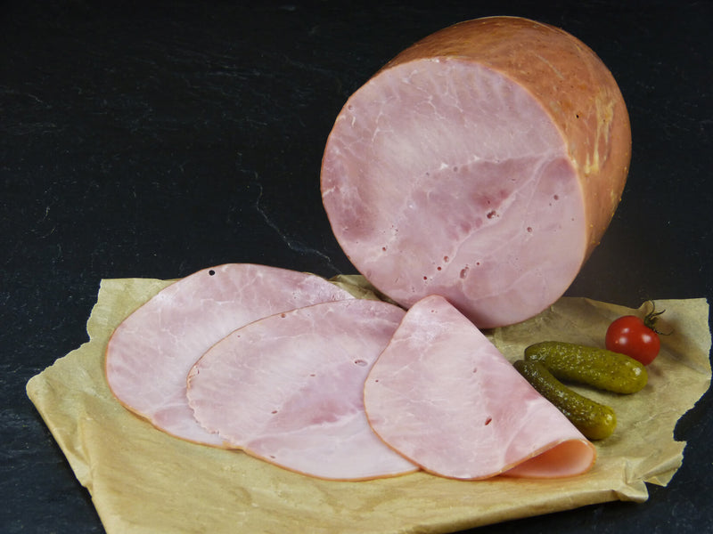 Gekochter Hinterschinken Kochschinken geschnitten und ein halbes Stück mit Essiggurken und Tomaten auf Schieferhintergrund