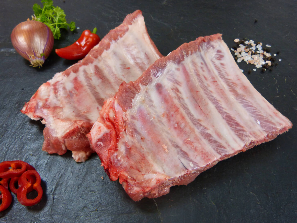 Spareribs von der Brustspitze auf Schieferhintergrund angerichtet mit Chili Zwiebel Kräutern und groben Salz und Pfefferkörnern