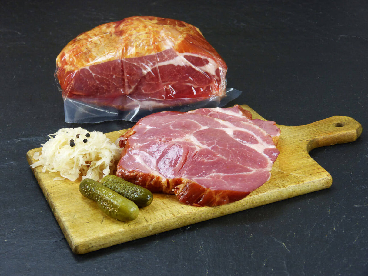 Kasseler Nackenbraten auf Holzbrett angerichtet mit Essiggurken und Krautsalat auf Schieferhintergrund