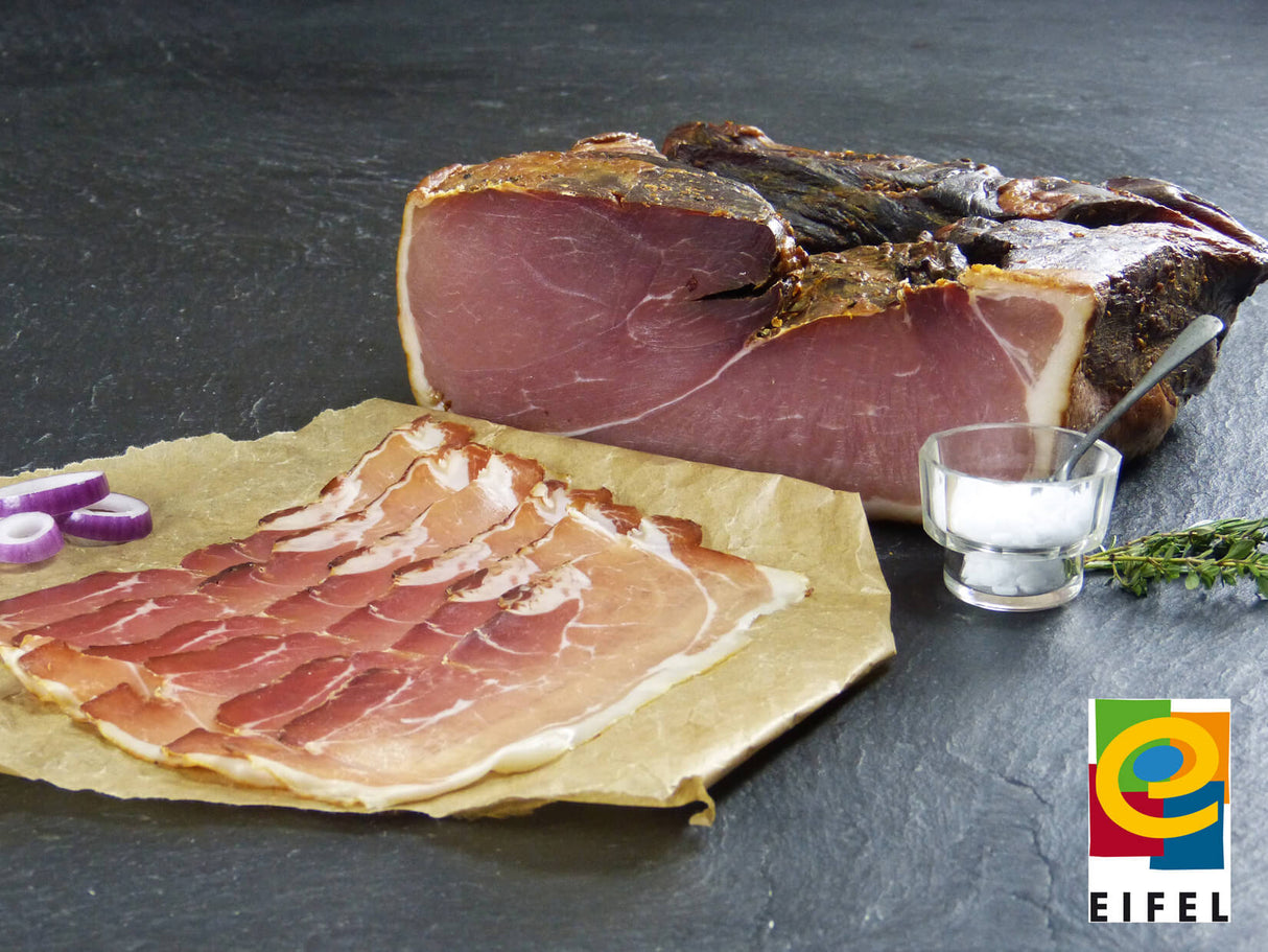 Bauernschinken vom EIFEL Schwein aufgeschnitten auf Backpapier angerichtet mit Zwiebelscheiben auf Schieferhintergrund