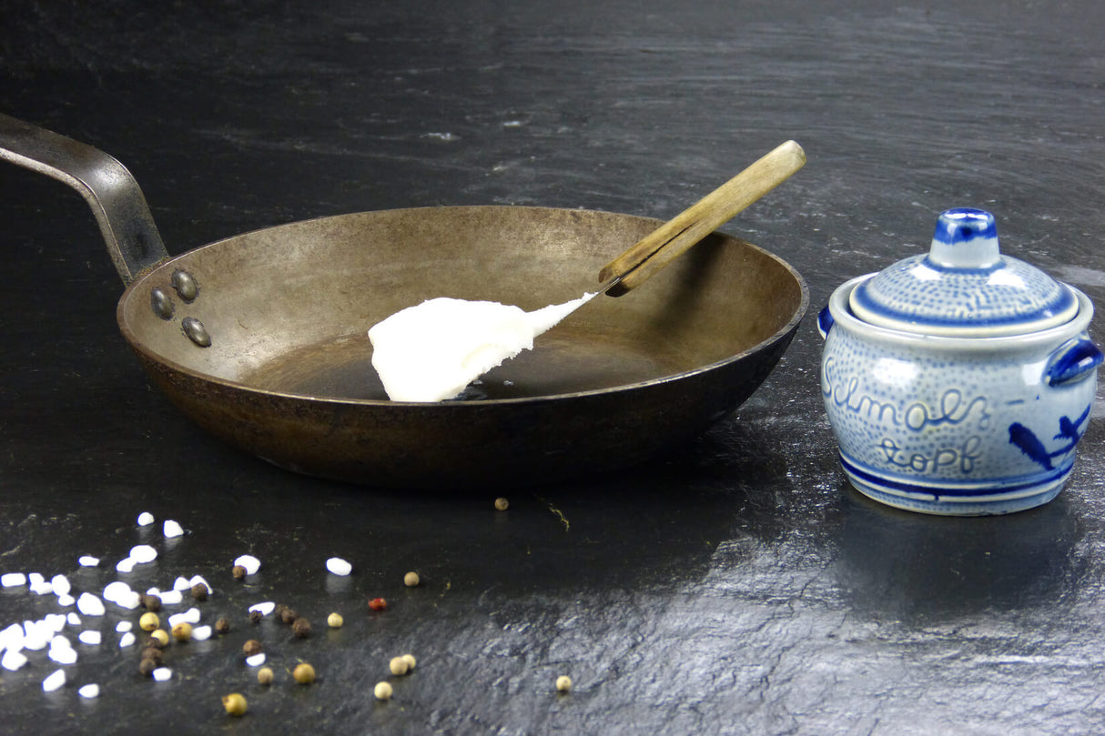 Schmalz vom EIFEL Schwein angerichtet auf Messer und Gusspfanne auf Schieferhintergrund mit groben Salz und Pfefferkörnern und Schmalztopf