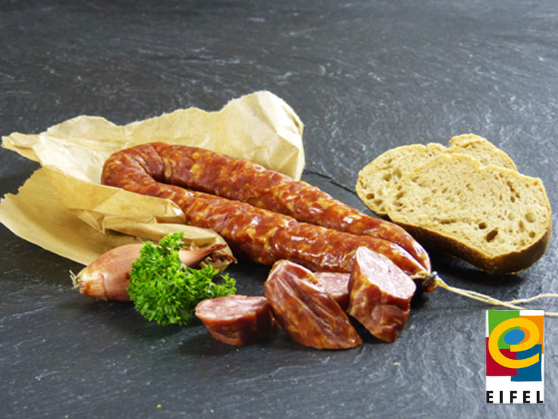 Luftgetrocknete Mettwurst vom EIFEL Schwein angerichtet auf Backpapier mit einer kleinen Zwiebel, Petersilie und Baguettescheiben auf Schieferhintergrund