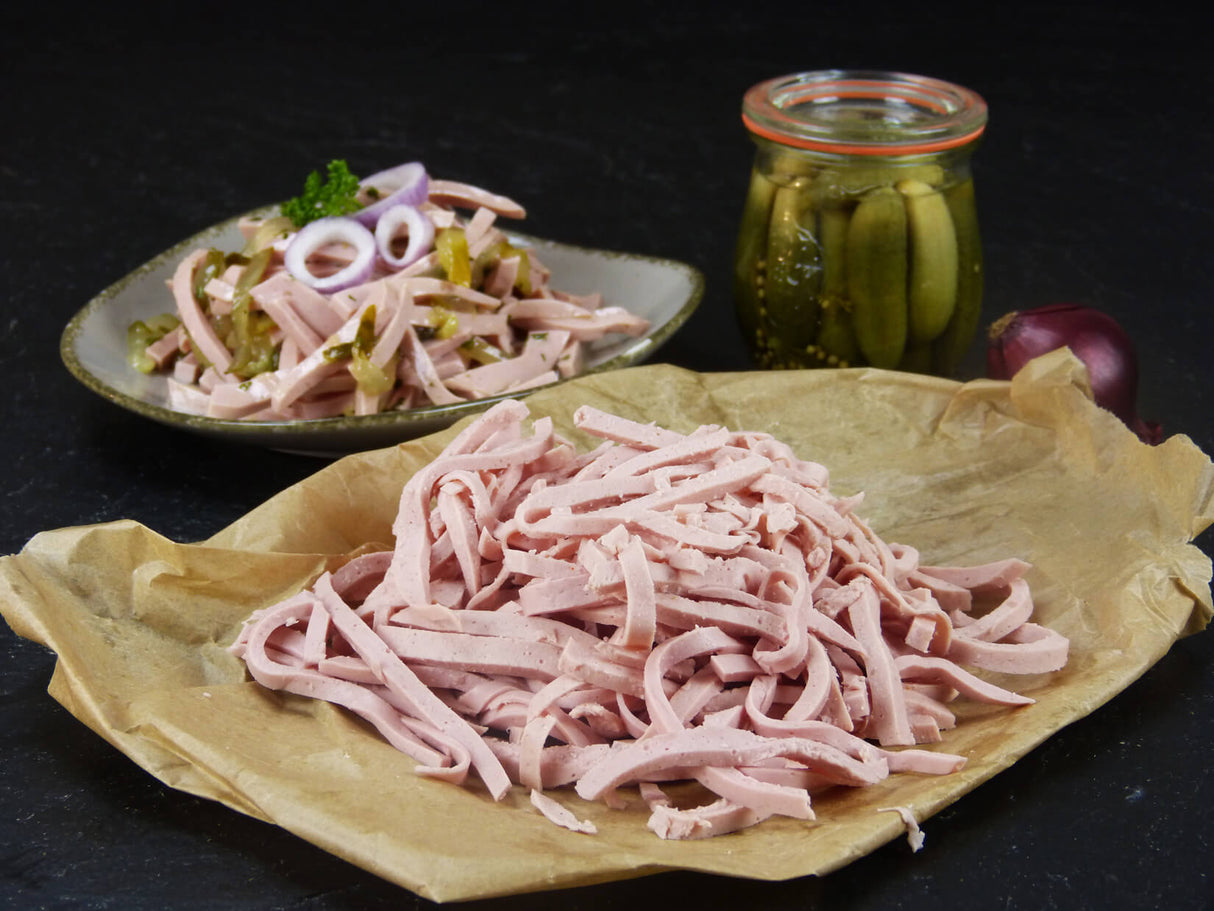 Fleischwurst in Streifen in einer Schüssel mit Zwiebeln Petersilie und Gewürzgurken in Scheiben neben einem Gewürzgurkenglas auf Schieferhindergrund