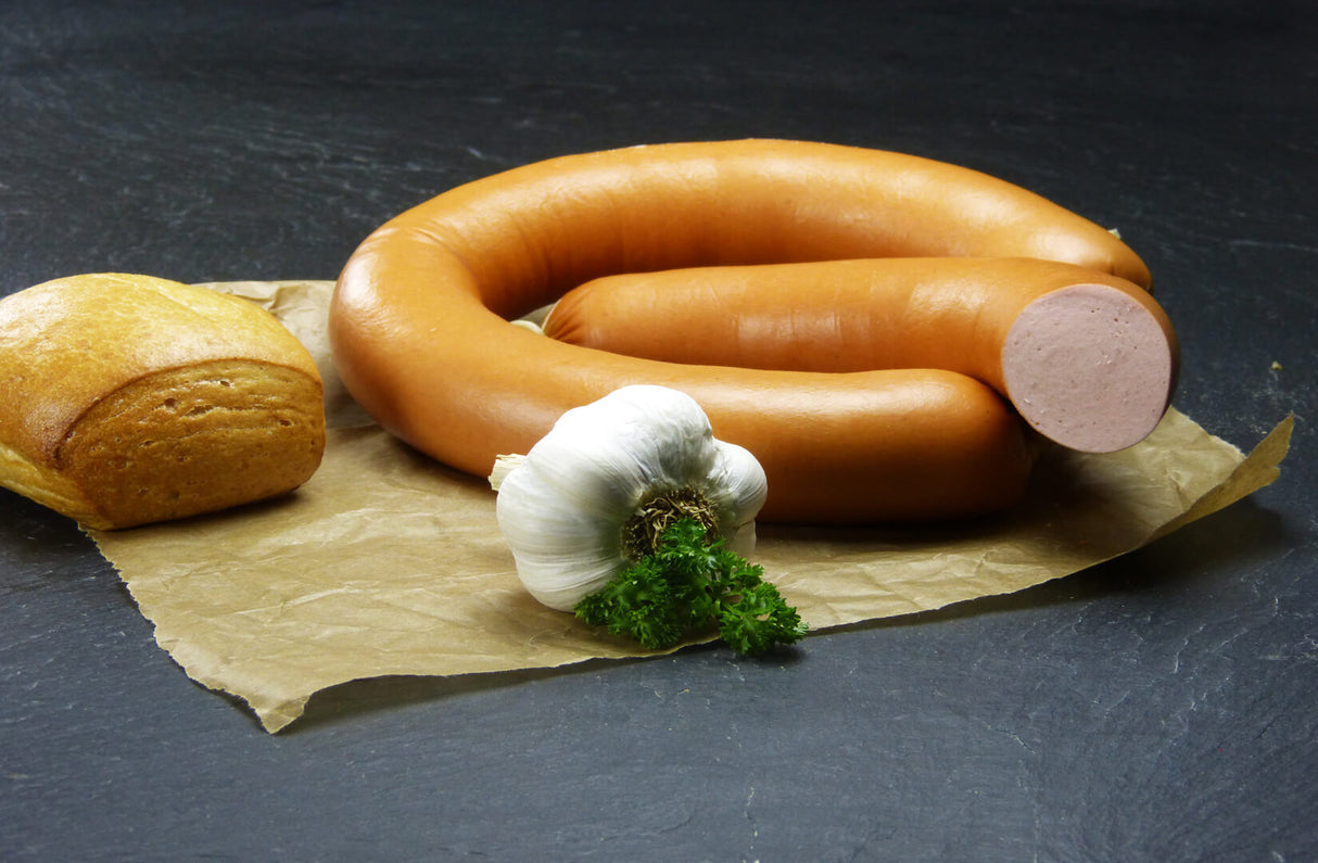 Fleischwurst im Ring mit Knoblauch halber Ring vom EIFEL Schwein angerichtet auf Backpapier mit einer Knoblauchknolle Petersilie und einem Brötchen auf Schieferhintergrund
