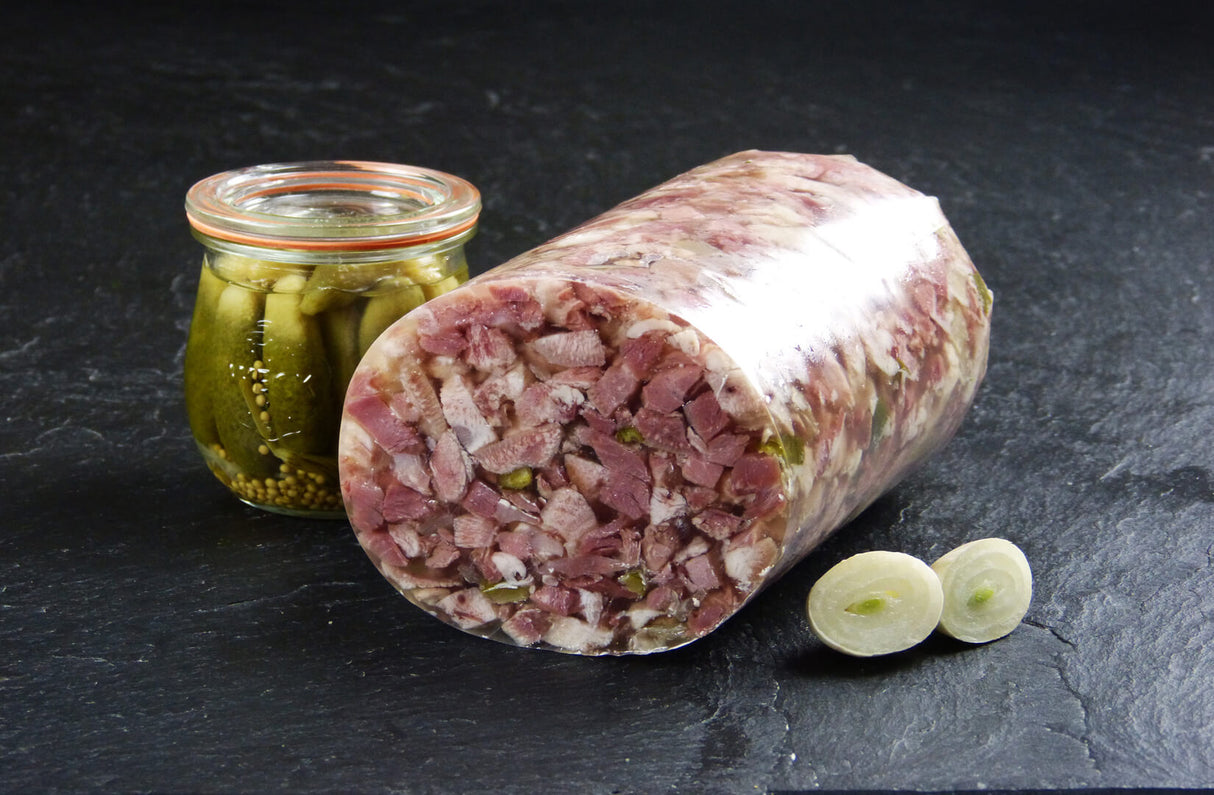 Schweinskopfsülze mit Gurken geschnitten mit einer Zwiebel und einem Gewürzgurkenglas auf Schieferhintergrund