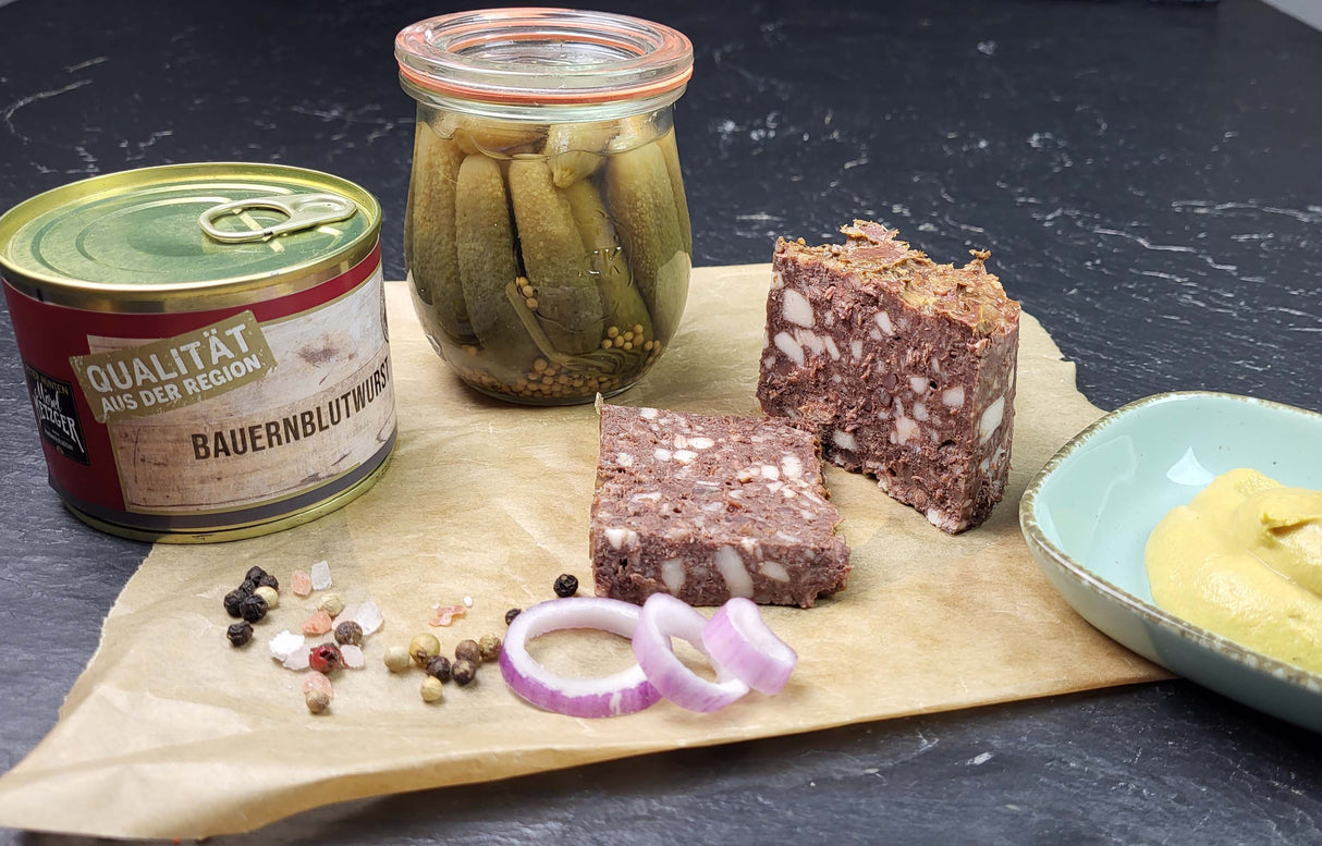 Bauernblutwurst 200g Dose vom EIFEL Schwein angerichtet auf Backpapier mit groben Salz und Pfefferkörnern roten Zwiebelscheiben einem kleinen Gewürzgurkenglas und einer kleinen Schale Senf auf Schieferhintergrund