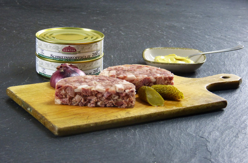 Schweinskopfsülze auf Holzbrett angerichtet mit roter Zwiebel und Essiggurke auf Schieferhintergrund mit einer kleinen Schale Senf