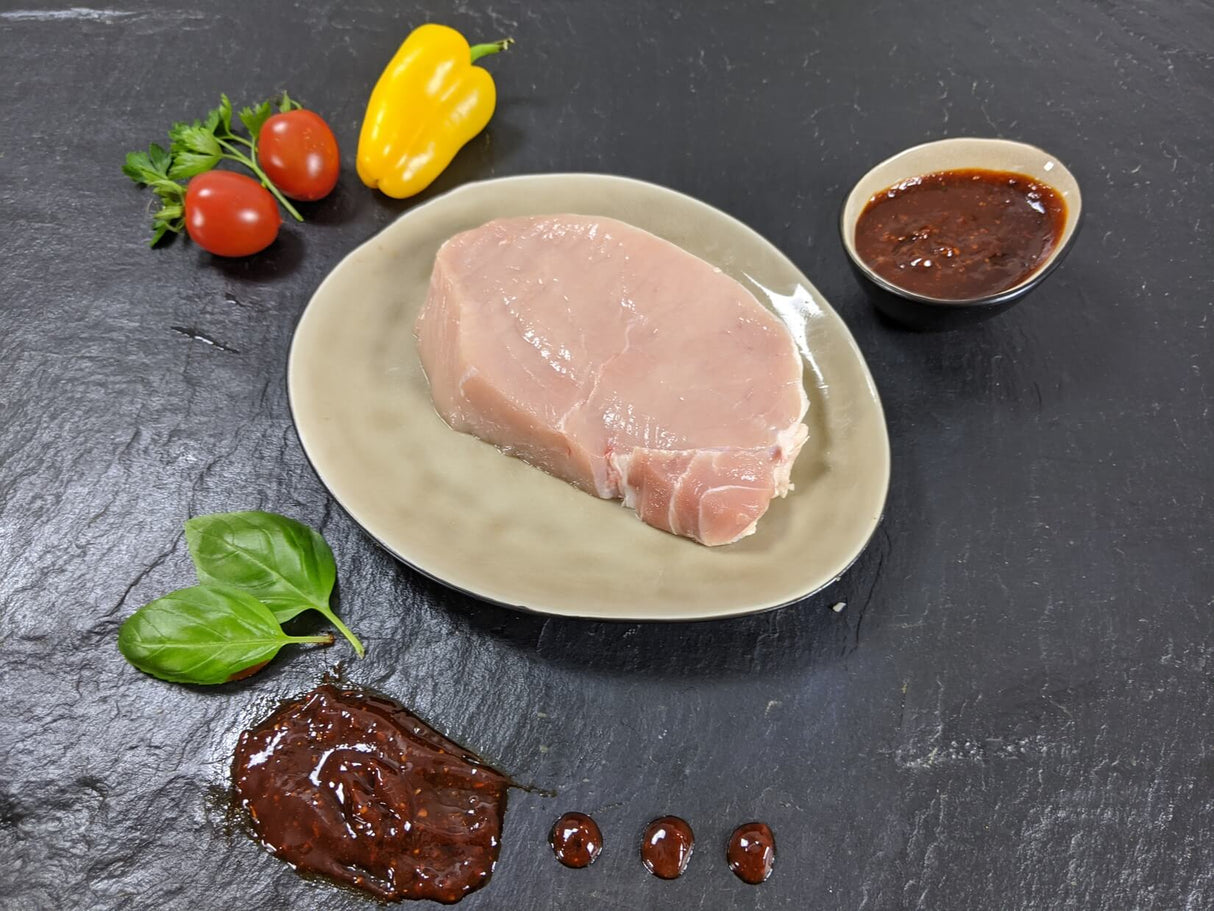 Schweinerückensteak Barbecue auf Teller angerichtet mit einer Schale Barbecue Marinade Tomaten Paprika und Kräutern auf Schieferhintergrund