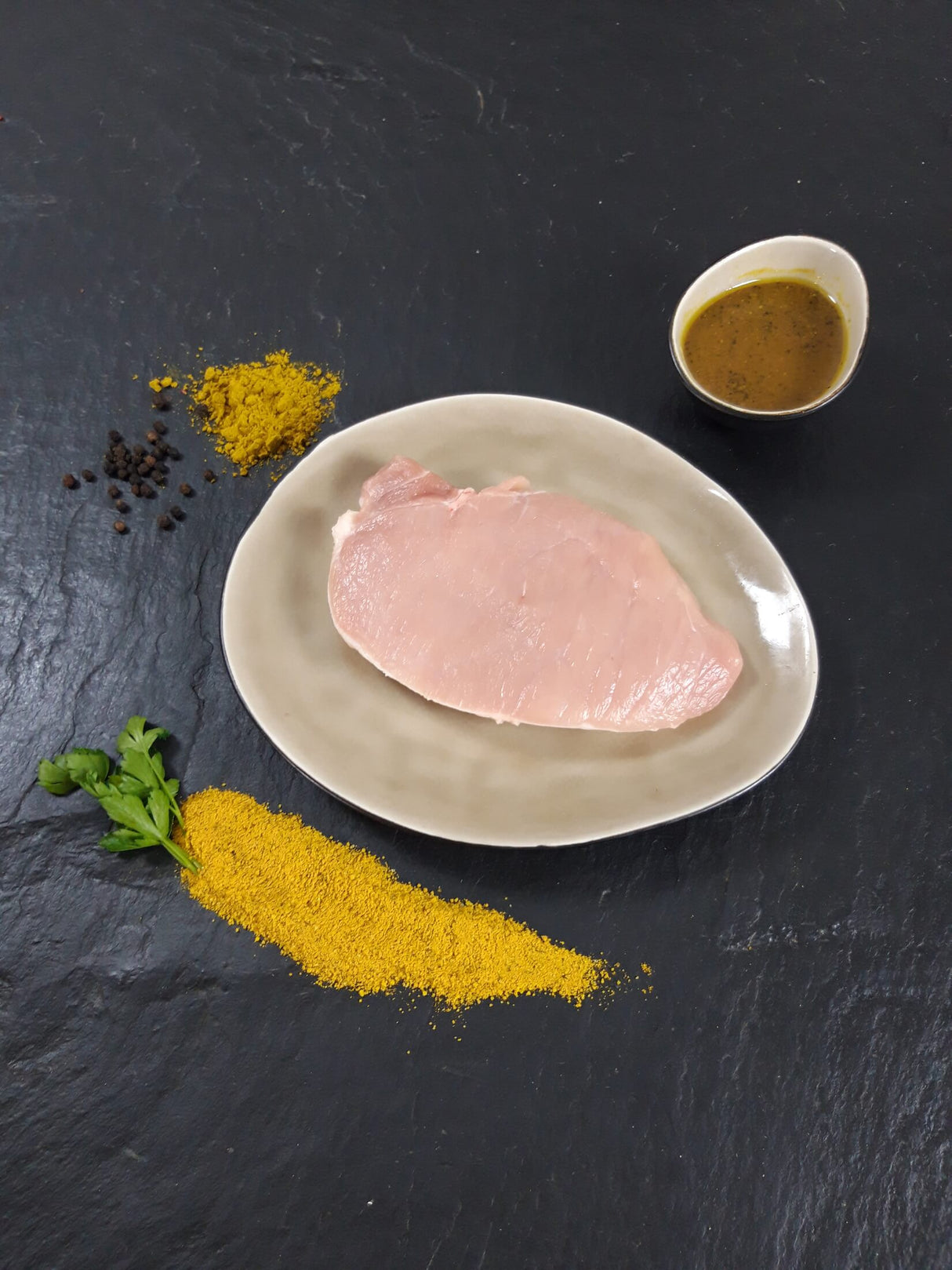 Schweinerückensteak Bombay auf Teller angerichtet mit einer Schale Bombay Marinade Gewürzen und Kräutern auf Schieferhintergrund