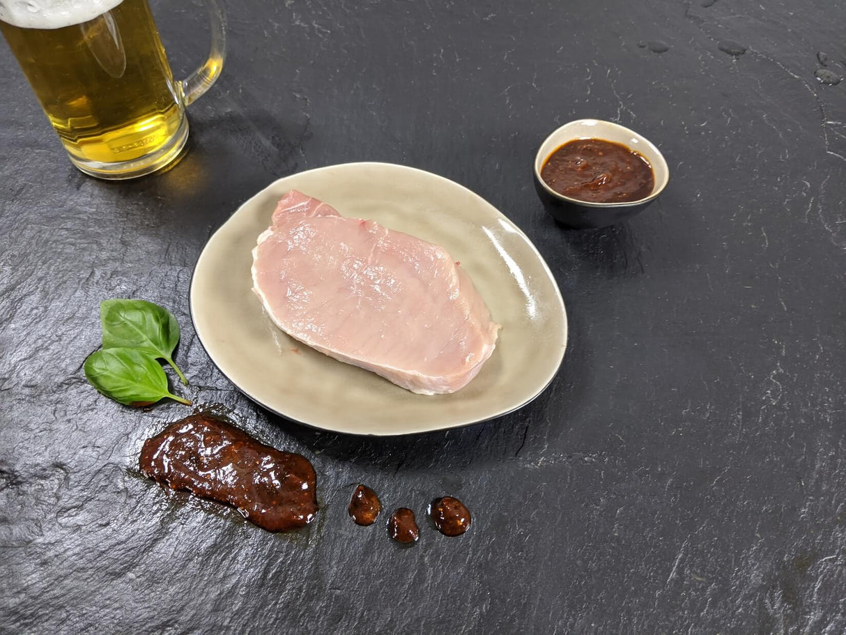 Schweinerückensteak Braumeister auf Teller angerichtet mit einer Schale Braumeister Marinade Kräutern und einem Glas Bier auf Schieferhintergrund