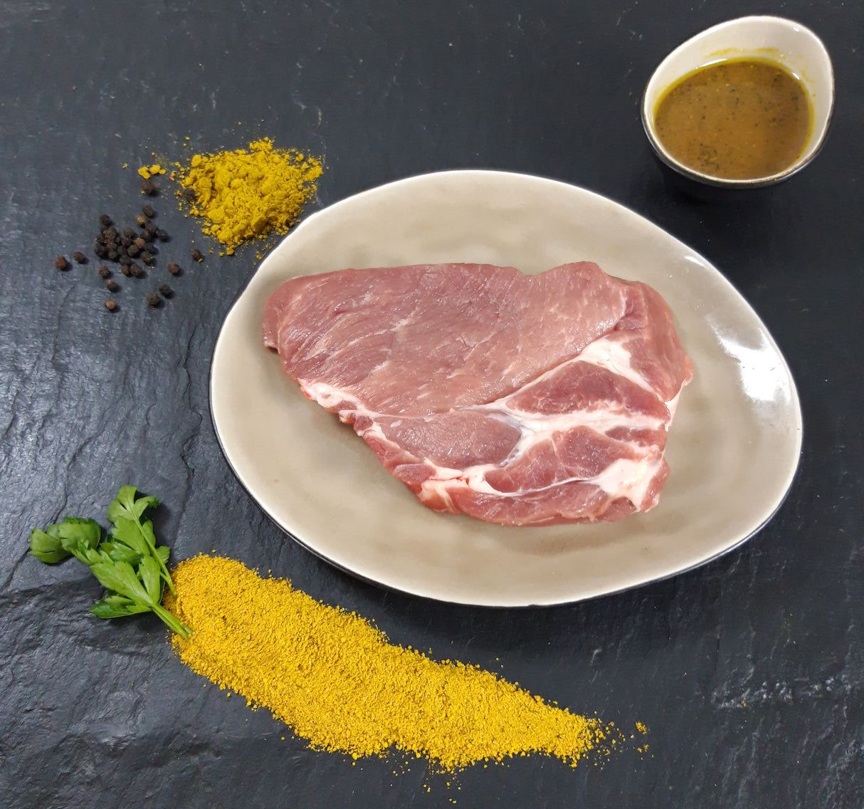 Schweinenackensteak Bombay auf Teller angerichtet mit einer Schale Marinade Kräutern und Currypulver auf Schieferhintergrund