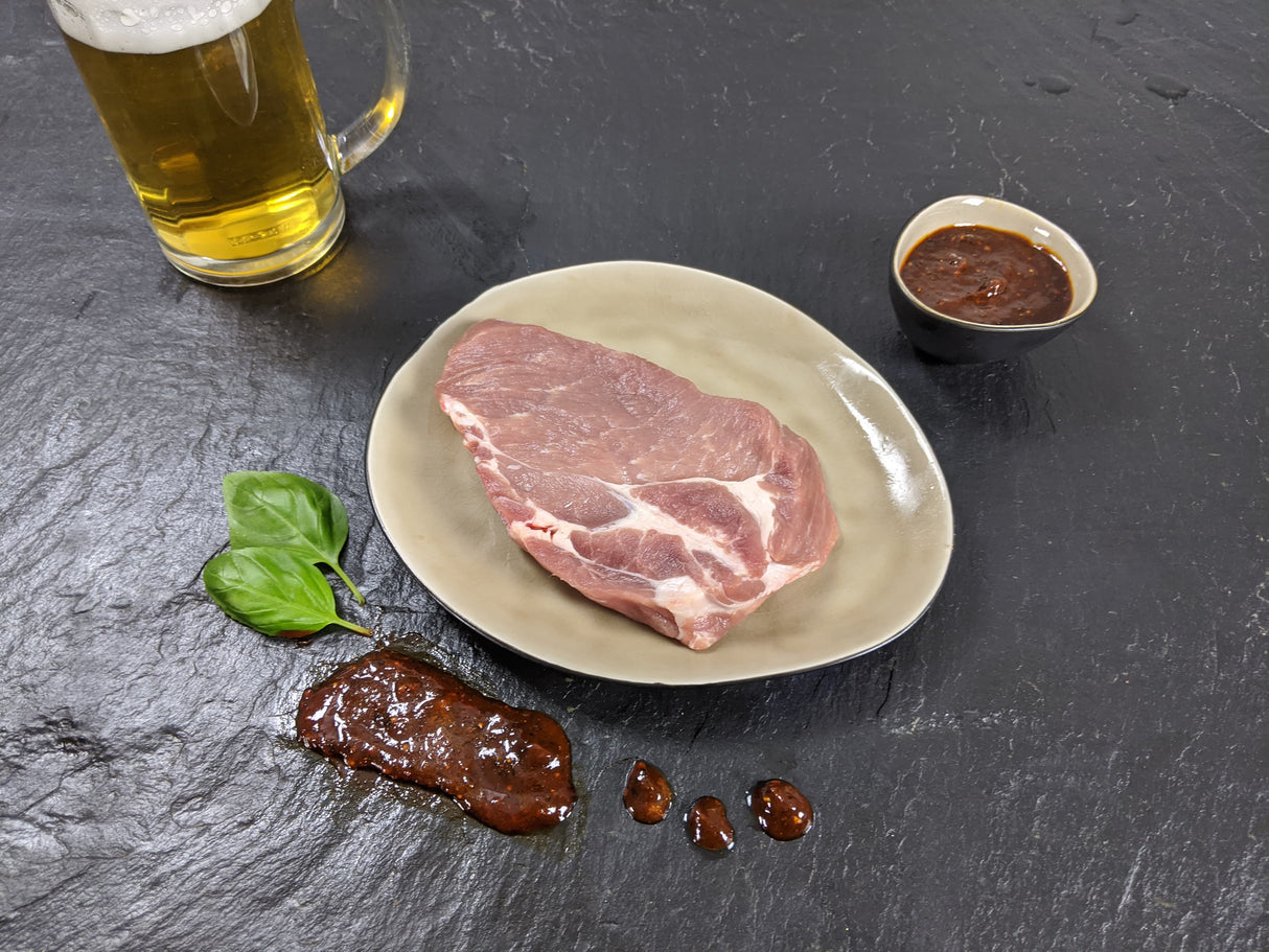 Schweinenackensteak Braumeister auf Teller mit einer Schale Marinade Kräutern und einem Glas Bier auf Schieferhintergrund