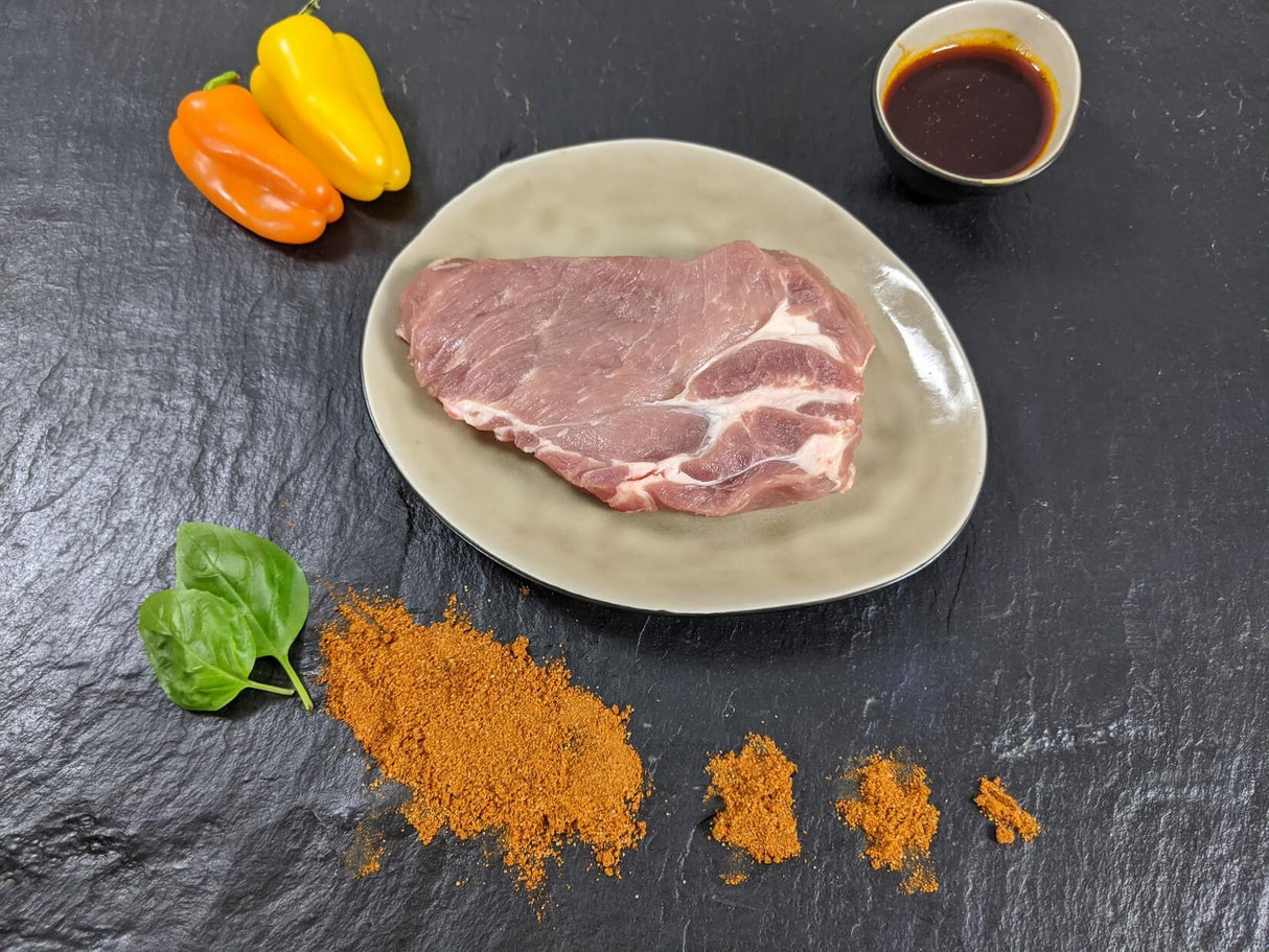 Schweinebauchscheibe Paprika auf Teller angerichtet mit einer Schale Marinade Paprika Kräutern und Paprikapulver auf Schieferhintergrund
