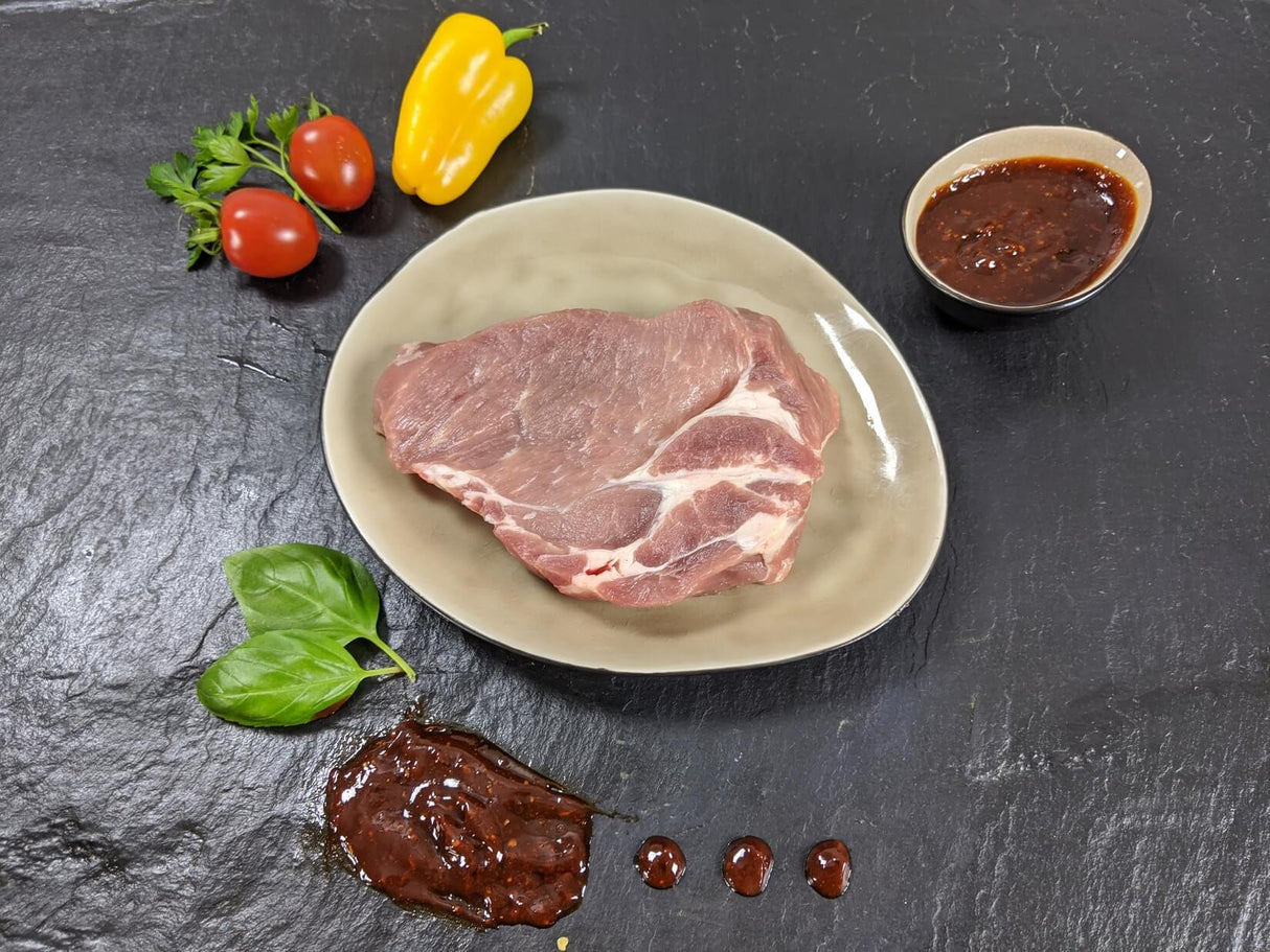 Schweinebauchscheibe Barbecue auf Teller angerichtet mit einer Schale Marinade Tomaten Paprika und Kräutern auf Schieferhintergrund