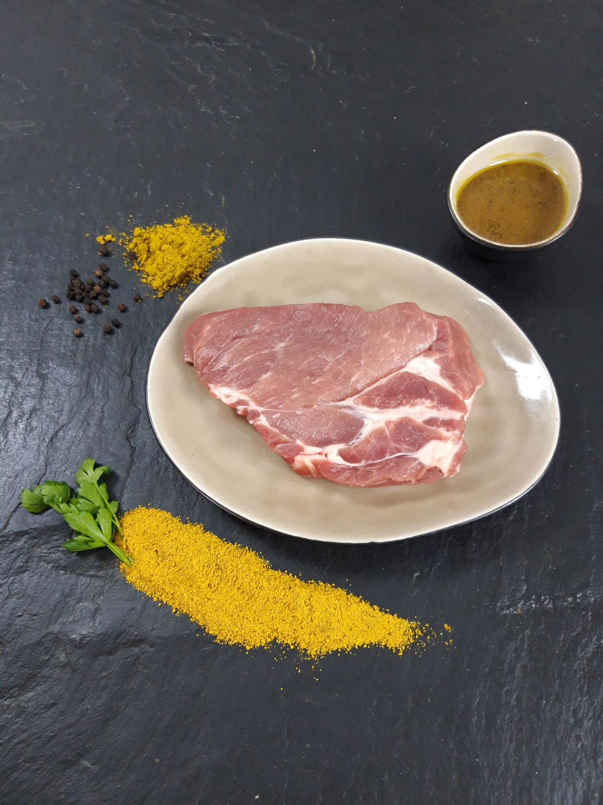 Schweinebauchscheibe Bombay auf Teller mit einer Schale Marinade Kräutern und Currypulver auf Schieferhintergrund