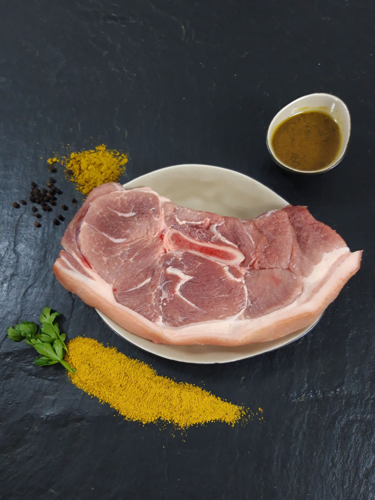 Holzfällersteak Bombay auf Teller angerichtet mit einer Schale Marinade Kräutern und Currypulver auf Schieferhintergrund